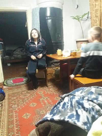 The image captures an interior scene. A young woman in a dark tracksuit sits on a chair near a large, dark oven or stove. A red patterned rug covers the floor. A person wearing gray and black stands at a wooden table. The location is possibly in a home or living space within the rural district of Borodulikha, Kazakhstan. The room is cluttered with objects, and an open doorway reveals a dimly lit space.