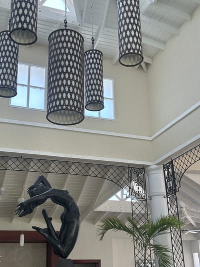 The image shows a sculpture of a woman in a contorted position, inside an open-air building in Punta Cana, Dominican Republic, at the coordinates 18.723 latitude and -68.465 longitude. The sculpture is made of a dark-colored material, possibly bronze. The woman’s arms are raised above her head and her legs are bent at the knees. The sculpture is standing on a pedestal, located in the center of the image. The building is designed with white walls, a wooden beamed ceiling, and white pillars. There are four hanging, rectangular, lattice-style lamps in the building. The building has a large open section with a lattice-style window and a palm tree. The building is a large, open space with a high ceiling. The image was taken from a slightly elevated angle, looking down at the sculpture.