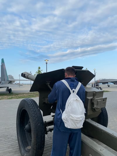 In Arablyar, Russia, a person stands facing away from the camera, examining a military artillery piece. The individual is wearing a dark blue tracksuit and a white corduroy backpack. The artillery piece is large and green, with a thick tire visible on the left side of the frame. In the background, two fighter jets are on display, one on the left with its tail fin showing and another further back on the right. The scene appears to be an outdoor exhibit, possibly a museum or memorial, with a paved walkway and a cloudy sky overhead.