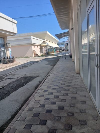 The image depicts a scene on a sunny day in the city of Gulistan, Uzbekistan, likely in a commercial or market area. In the foreground, a paved walkway is visible, featuring a patterned arrangement of hexagonal and square tiles in varying shades of gray and brown. To the left of the walkway is a paved road with a worn asphalt surface, exhibiting cracks and patches of gravel. On the right side of the walkway, a modern building with large glass windows and white frames runs parallel to the path. A white awning extends over the sidewalk in front of this building.

Further into the background, another building with a light-colored facade and a distinctive angled roofline is situated. This structure has an outdoor air conditioning unit mounted on its wall. Adjacent to this building, a large cylindrical water tank, possibly elevated on a metal frame, is visible. Beyond these structures, a low-rise building with multiple windows and awnings can be seen. The sky is clear and blue, with some power lines crisscrossing the upper portion of the frame.

The scene appears quiet, with no people prominently featured in the immediate vicinity. A single black chair is placed on the walkway, and a stack of chairs is visible near the entrance of a building to the left. The overall impression is of a functional outdoor space, possibly a service area or an entrance to shops or businesses.