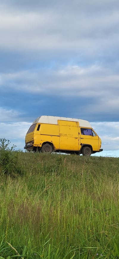 A yellow and white van is parked on a grassy hill under a cloudy sky. The van is oriented perpendicular to the viewer, with its front facing right. The van is weathered and appears to be an older model. The grass in the foreground is tall and green, with some dry stalks mixed in. The sky is filled with large, grey and white clouds, with patches of blue visible. The scene appears to be in a rural or undeveloped area, possibly a field or roadside in Hetmanivka, Ukraine. There are no people visible in the image.
