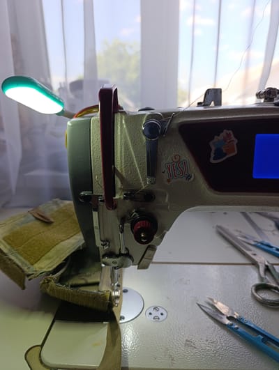 A sewing machine is positioned on a table, illuminated by a bright green LED light. The machine, predominantly cream-colored with maroon accents, features various metal components and a red adjustment knob. A single thread is visible, extending from the top of the machine. Attached to the machine's body are two stickers: one depicting potted plants, and another with the word "YES!" in colorful letters. In the foreground, a piece of olive-drab fabric with Velcro is being sewn, with another piece of fabric already attached to the sewing machine. To the right, a pair of small blue-handled scissors and larger, metal-handled scissors lie on the table. In the background, sheer white curtains are drawn, with a glimpse of greenery visible through them. The overall scene suggests a workspace dedicated to sewing, with tools and materials neatly arranged. The location of Filippenkov, Russia, is noted, but the image itself provides no direct evidence to confirm or deny this specific location.