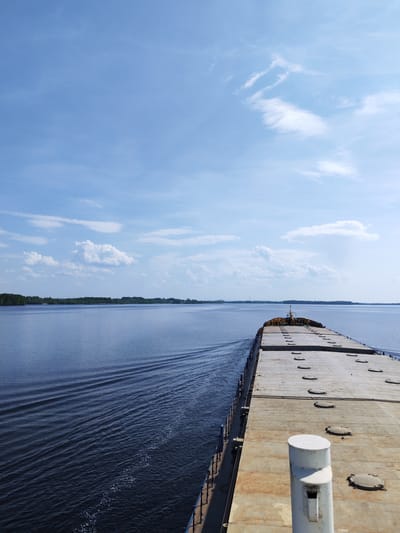 The image depicts a wide-angle shot from the deck of a barge moving on a body of water. The deck is concrete with numerous access hatches and is framed by a railing on the left. The water is rippled with the wake of the barge visible. In the distance, the shoreline is marked by trees. The sky is blue with scattered clouds. This view appears to be on the Rybinsk Reservoir in the city of Bessolovo, Russia.
