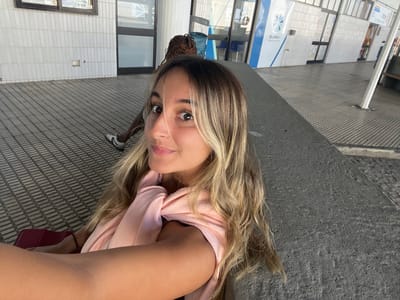 The image displays a young Caucasian female taking a selfie at the BusWay bus station in Albergaria-a-Velha, Portugal. She has long, wavy blonde-highlighted hair, dark eyes, and wears a light pink garment draped over her shoulders. Her right arm is extended, holding the camera, with a dark bracelet visible on her wrist. A dark red object, possibly luggage, is partially visible to her left. In the background, to the left, a person of dark complexion with braided hair is partially visible, showing a leg and a white sneaker with a dark stripe. The setting is an outdoor public transit area featuring a dark concrete ledge in the foreground, dark gray rectangular floor tiles, and a building with light-colored horizontal wall tiles. Dark-framed windows and doors are present on the building's facade. A prominent blue sign displaying "BusWay" and a white circular logo identifies the location as a bus terminal.