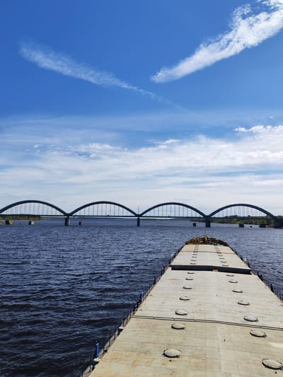 A long, flat-topped barge stretches into the water toward a large arched bridge over the Volga River, under a bright blue, partly cloudy sky in Volga, Russia. The barge is grey and textured, with numerous circular hatches covering its surface. The water is dark and rippled, reflecting the blue sky. The bridge is a series of arches, with power lines visible on one side. The sky is a vibrant blue with scattered white clouds. The scene appears to be captured from an elevated position on the barge itself.