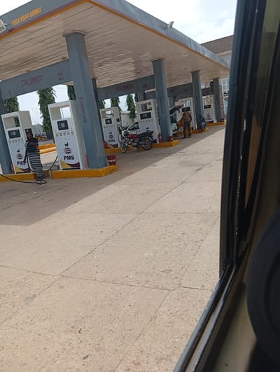 This media file captures a view of a gas station on Yola Road, Gombe, Nigeria. The scene is dominated by several gas pumps under a covered canopy. A woman in a striped dress stands near one of the pumps, with a fuel hose extending from it. Several other people are visible near the pumps, some attending to vehicles, while others appear to be waiting. A motorcycle is parked near one of the pumps. The ground is paved with concrete. The sky above is partly cloudy, suggesting it's daytime. The gas pumps are labeled with "PUMP" and numbers, along with branding that includes "MRS" and "PMS". There are also "NO SMOKING" signs visible. The overall impression is of a typical operational day at a Nigerian gas station.