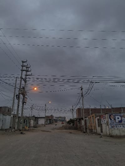 This image displays an unpaved residential street in Nuevo Chimbote, Peru, observed under a dark, overcast sky during dusk or nighttime. The street is composed of light brown/grey dirt and debris, extending into the distance and appearing to narrow. Dominating the foreground and midground are numerous concrete utility poles lining both sides of the thoroughfare, heavily laden with intricate networks of overhead electrical and communication wires. Two streetlights are illuminated with an orange glow, one prominent on the left and another further down the street on the right.
Structures bordering the street consist primarily of single-story buildings or dwellings, appearing rudimentary or unfinished, constructed from various materials including concrete blocks and possibly wood or corrugated metal. On the immediate right, one building's facade is adorned with multiple political posters, displaying faces, the letter "P," and numerical sequences such as "P123." Further down the street on the right, a larger, uncompleted brick structure is visible.
In the mid-distance on the dirt road, a black canine is present, standing near a pile of sand or gravel. A red vehicle, possibly a car or small truck, is discernible in the far distance on the road. No human figures or direct human interactions are visible. The overall environment suggests a developing urban or semi-urban area.