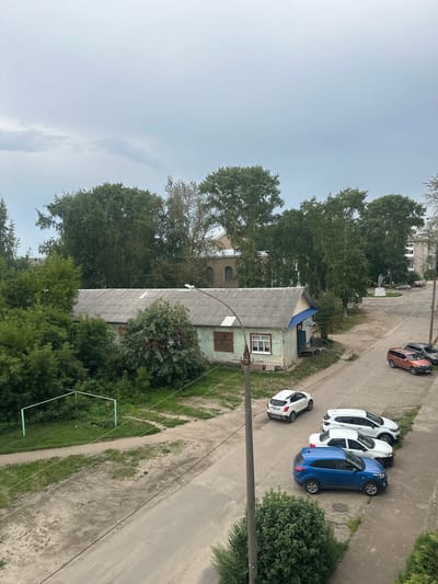 This aerial, vertical shot captures a residential scene in Kotlas, Russia, likely during daylight hours under an overcast sky. The image showcases a row of parked cars, including a blue SUV, two white hatchbacks, and an orange hatchback, all positioned on a paved area adjacent to a street. A small, dilapidated building with a grey corrugated roof and light green walls, featuring a single window and a smaller, older window with wooden shutters, is prominent. Beyond this building, dense green trees form a backdrop. In the distance, a larger building with multiple windows and a lighter colored facade is visible, along with what appears to be a statue. The scene suggests a quiet, ordinary day in a somewhat weathered urban or suburban environment. There is no visible text in the image.