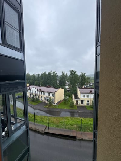This is a vertical, eye-level, exterior shot of a residential area in Близкое Кп, Russia. The weather appears overcast and possibly rainy, with wet asphalt visible on the road and sidewalks. The scene depicts several yellow apartment buildings with dark red roofs, surrounded by green grass and trees. The foreground features a modern building with large windows, reflecting the surrounding environment. Two identical apartment buildings, each with multiple windows and balconies, are prominently featured. One building appears closer to the foreground than the other. The overall setting suggests a quiet, suburban residential neighborhood. There is no visible text within the image.