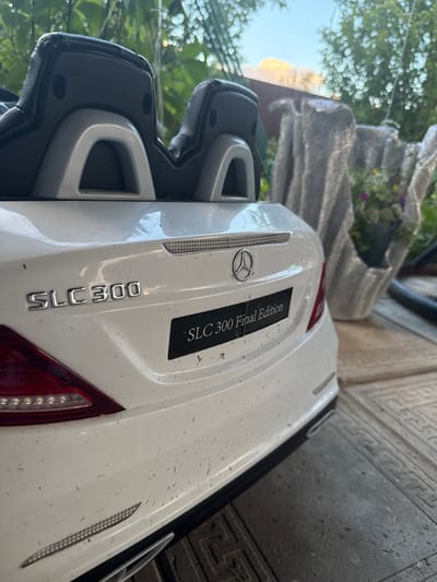 This media file shows a close-up, angled view of the rear of a white toy car, likely a Mercedes-Benz SLC 300 Final Edition, parked outdoors. The car is slightly dusty, with visible specks of dirt and debris on its surface. The iconic Mercedes-Benz star emblem is prominent on the rear panel. To the right of the emblem, a black sticker reads "SLC 300 Final Edition." Below this, the "SLC 300" badge is also visible in silver lettering. The car has two dark-colored seats and a white body.
In the background, out of focus, are elements suggesting an outdoor setting with greenery from trees, and possibly a light-colored stone or concrete structure, perhaps a planter. The lighting suggests it is daytime, with diffused sunlight. The overall scene appears to be a residential yard or driveway. No people are visible. The text visible on the car clearly identifies it as a "SLC 300" and a "Final Edition" model of what is presumably a children's ride-on toy.