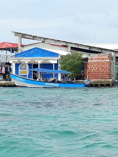 In the city of Unknown, Colombia, a blue and white boat named "JUANJO" is docked next to a blue building with white text reading "IGLESIA CRISTIANA PENTECOSTES MOVIMIENTO MISIONERO MUNDIAL" and a logo with a globe. The boat has a Yamaha outboard motor. Several people are seated on the boat. To the left of the building, laundry hangs on a clothesline. Adjacent to the blue building is a structure made of red bricks. The scene is situated on the water, which appears calm and turquoise. The sky is overcast.