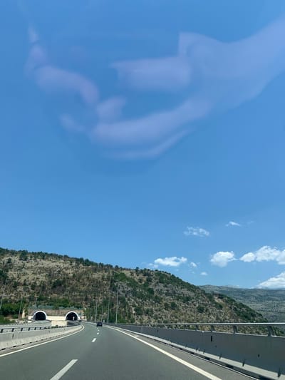 The image shows a highway in Gornje Mrke, Montenegro, leading towards two parallel tunnel entrances in a rocky, vegetated hillside. The highway has clear lane markings and concrete barriers along its sides. A black car is visible driving on the highway. The sky is blue with some light cloud formations.