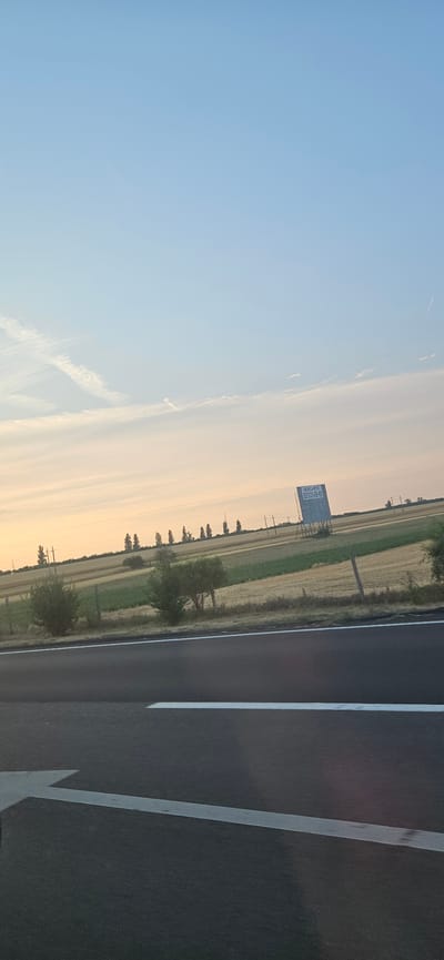 This is a photograph taken from a roadside, likely on a highway, looking out over a rural landscape at sunset or sunrise. The sky is a soft gradient of pale blue to warm orange. In the distance, a billboard with text that appears to be in Hungarian is visible, mounted on a metal frame. The text reads "BERELHEVÉTO" and a phone number "70/333-82-55". The landscape consists of fields with varying textures and colors, suggesting agricultural use, with rows of trees lining the horizon. Power lines and poles are also present. In the foreground, the asphalt road surface with white lane markings is clearly visible. No people are present in the image. The scene appears to be located in or near Székesfehérvár, Hungary, given the Hungarian text on the billboard.