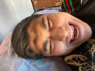 The image displays a close-up of a young male subject, positioned horizontally with his head rotated approximately 90 degrees clockwise relative to the viewer. This scene is located in the city of Mirzabashichek, Uzbekistan. The individual has dark, short hair and a medium skin tone. His eyes are dark and tightly squinted, exhibiting visible wrinkles at the outer corners. His mouth is wide open, revealing a full set of white teeth, pink gums, and the tongue. Faint facial hair is present on the upper lip and chin. He is attired in a dark garment featuring a distinct green and red striped pattern on the visible upper arm or shoulder. The subject lies on a pillow covered by a patterned pillowcase with hues of purple, blue, and pink. A dark blanket or fabric with lighter, possibly gold-toned, geometric patterns is visible beneath his head and partially over his body. The background consists of a plain, light-colored wall and a section of what appears to be worn wooden furniture or a door frame in the upper right quadrant. Strong, directional lighting illuminates the left side of the subject's face, casting portions of the right side into shadow. The facial expression suggests intense laughter or a strong vocalization.