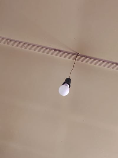 A single bare light bulb hangs from a brown wire attached to a ribbed strip on a tan ceiling in Sarıağaş, Kazakhstan. The light bulb is illuminated and white, with a black socket. The ceiling appears to be textured or painted, with some small dark specks visible. The lighting is dim.