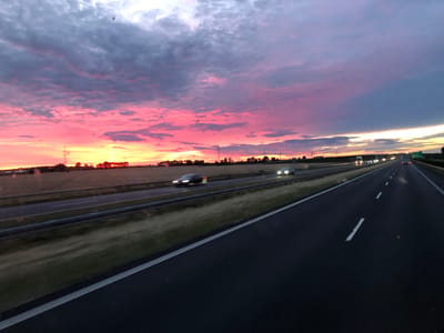 The image depicts a highway scene at dusk in Antoninek, Poland, under a dramatic sky filled with pink and purple clouds. The highway stretches into the distance, with vehicles in motion, their headlights illuminated. On the left side of the highway, a field of crops is visible, bordered by trees and power lines. The overall impression is one of a dynamic and colorful transition from day to night, captured from a moving vehicle on a highway.