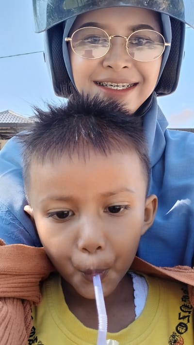 In Matangbaroh, Indonesia, a selfie captures two individuals, likely a mother and child, on a motorcycle. The woman, wearing a blue hijab and round gold-rimmed glasses, smiles, revealing braces. Her helmet is visible, along with a portion of a building's roof in the background. In the foreground, a young child, facing forward, is drinking from a purple straw. The child's hair is styled upward, and they are wearing a yellow shirt with a brown blanket draped over their shoulders. The scene suggests a sunny day, with a clear sky.