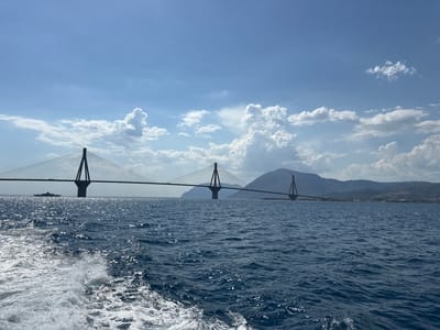 The image captures a wide-angle view of a large cable-stayed bridge spanning a body of water. The bridge is the dominant object in the scene, with its tall pylons and suspended roadway stretching across the horizon. In the foreground, the foamy wake of a boat indicates movement, suggesting the perspective is from a vessel. The water is a deep blue, with sunlight glinting off its surface, creating a dynamic and textured appearance.
In the distance, a ferry boat is visible near the base of one of the bridge's pylons, adding a sense of scale. The background features rolling hills and mountains, partially obscured by a hazy atmosphere. A cluster of wind turbines can be seen on the hillside in the right portion of the image, suggesting a wind farm. The sky is a vibrant blue, populated with scattered, fluffy white clouds that are characteristic of a fair weather day. The lighting suggests it is daytime, likely late morning or early afternoon, given the brightness and the angle of the sun.
The overall mood is serene and majestic, highlighting the impressive engineering of the bridge against a natural landscape. The location is the Municipal Unit of Rio, Greece, which is known for the Rio–Antirrio Bridge, a prominent example of a cable-stayed bridge. There is no visible text in the image.