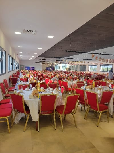 Rows of tables with red chairs fill a large hall, ready for an event. White tablecloths cover each table, set with plates, glasses, and bottles. Decorations hang from the ceiling. The floor is light brown, and the walls are white, with windows along one side. The ceiling is a combination of white and dark gray panels. The space is brightly lit by ceiling lights.