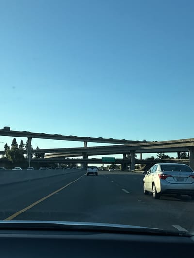 The image shows a highway with multiple overpasses. A silver car is visible in the right lane, with the license plate "8JGV889". A yellow line separates lanes. A sign indicates "Santa Clara Street". The location is likely in or near San Jose, United States of America. There is traffic in the distance. The sky is clear and blue.