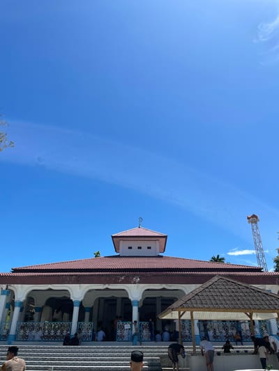 The image depicts a building with a red-tiled roof and white arched openings, possibly a mosque or a religious structure, under a clear blue sky. People are visible inside the building, some appearing to be engaged in prayer or other religious activities. A metal tower structure with speakers is also visible against the sky in the distance. The structure is located in or near Pasar Paloh, Indonesia, based on the location context.