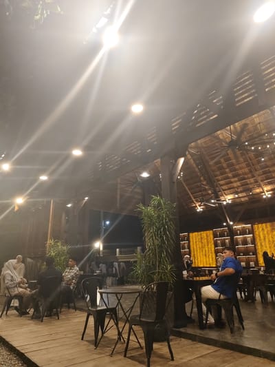 The image depicts an evening scene at an outdoor cafe. The setting is rustic, with a wooden roof structure, exposed beams, and warm lighting. Several people are seated at tables, engaged in conversation or enjoying their time at the cafe. In the foreground, a man in a blue t-shirt and white pants is seated at a table. To the left, a group of people, including two women in headscarves, are seated on a wooden deck. Further in the background, more patrons are visible, some standing and others seated. The cafe appears to be in a tropical or warm climate, suggested by the foliage and open-air design. The lighting indicates it is nighttime, and the overall atmosphere seems relaxed and social. There are decorative elements such as shelving with framed items on the wall and large ceiling fans, contributing to the ambiance. The scene is located in Bireuenmeunasahdayah, Indonesia.