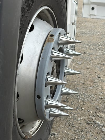 A close-up, eye-level shot captures the wheel of a truck or heavy-duty vehicle, featuring a distinctive spiked hubcap, set against a backdrop of gravel ground in Kamyennoye, Russia.
The primary subject is a large black tire, showing visible tread patterns and some unreadable text on its sidewall. It is mounted on a silver-painted metal wheel, which appears somewhat worn. Over the center of the wheel, a prominent grey, circular metal ring is attached, serving as a decorative or protective cover. This ring is embellished with eight shiny, chrome-like conical spikes, evenly spaced around its circumference. These spikes are reflective, catching the light and showing subtle striations on their surfaces. The grey ring itself exhibits some signs of age, with patches of rust and general wear. Beneath the spiked ring, part of a lighter silver hubcap is visible, with several cutouts.
The background consists of a rough, light brown and grey gravel surface, suggesting an outdoor, possibly industrial or unpaved parking area. To the left, a sliver of a white vehicle's bodywork or chassis is visible, indicating the vehicle to which the wheel belongs. The scene is illuminated by natural daylight, casting subtle shadows and highlighting the reflective qualities of the spikes. No activity is taking place, and the image presents a static view of the vehicle component.
