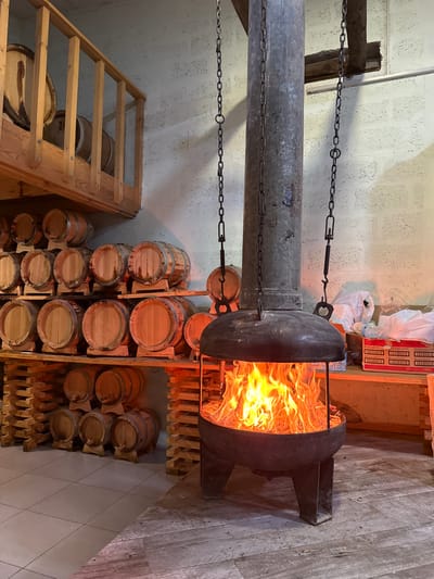 This image shows an indoor scene with wooden barrels and a fireplace. The barrels are stacked on shelves and on the floor. The fireplace is in the foreground and has a fire burning inside. There are also some boxes and bags in the background. The overall impression is one of a rustic and warm environment.

There is no indication of the location being in the city of Sisian, Armenia.
