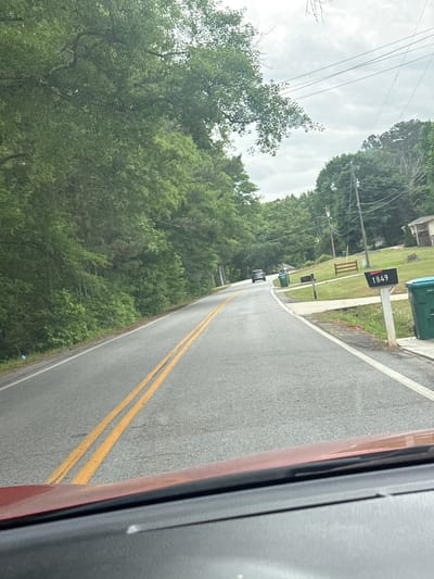 A street scene in Mountain Park, United States of America, captured from a moving vehicle. The view is from the passenger side, with a portion of the car frame visible on the right. A paved road stretches ahead, marked with double yellow lines. The roadside features mailboxes displaying house numbers; one visible mailbox shows "1849". Green trees are abundant on the left, and power lines crisscross the sky. A car is visible driving on the road in the distance. The sky is overcast.