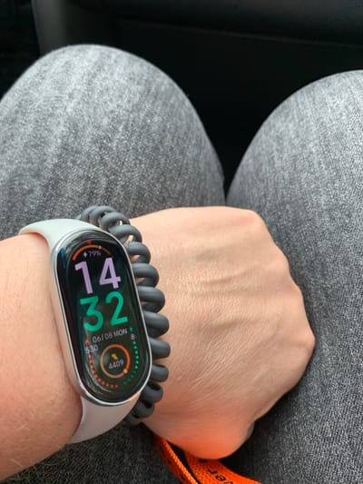 An overhead shot shows a person's left wrist wearing a smartwatch with a gray band and a coiled black strap extender. The smartwatch display shows the time as 14:32, a date of 06/08 MON, a battery level of 79%, and step count data. The wrist is resting on the person's lap, which is covered by gray jeans. The background is dark and out of focus. There are no discernible elements that would suggest a specific location within Сваткаўскі сельскі Савет, Belarus.