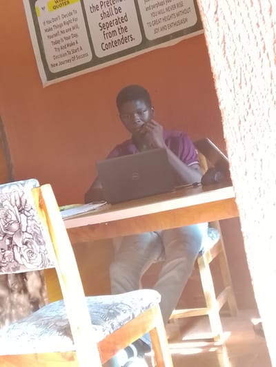 A young man is seated at a wooden desk, focused on a Dell laptop. He is dressed in a purple polo shirt and light-colored trousers. His left hand is raised to his face, seemingly in thought. On the desk to his right is a black computer mouse, along with some papers.
Behind him, the wall is a brownish-orange color and adorned with motivational posters. One poster features the phrase "the Pretenders shall be separated from the contenders" while another states "You will never rise to great heights without joy and enthusiasm."
The scene appears to be indoors, possibly a home office or study area. The foreground features a chair with a patterned seat cushion, and sunlight streams in from the right side of the frame. This location is Oke Ogun, Nigeria.