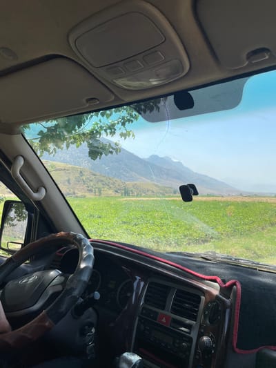 The image shows the interior of a vehicle looking out through the windshield. The view from the car reveals a sunny landscape with green fields, hills, and mountains in the distance. The foreground includes the steering wheel and dashboard of the car, with a red trim around the dashboard. A tree with green leaves is visible hanging over the windshield, partially obscuring the view. There are also some objects attached to the windshield, including what appears to be a dashcam or GPS device. The overall impression is of a scenic drive through a rural area. The location is identified as being in the city of Khaidarkan, Kyrgyzstan.