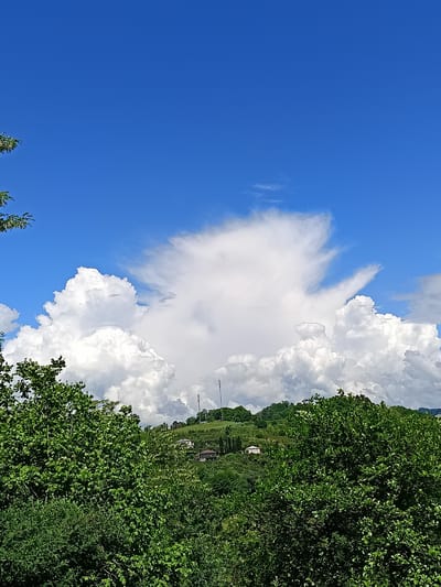 The image shows a landscape view with a large, towering cumulonimbus cloud formation in the center, dominating the sky. The sky is predominantly blue with some thinner clouds interspersed with the cumulonimbus. In the midground, a green, tree-covered hill can be seen with two visible white buildings and at least two communication towers atop the hill. In the foreground, lush green foliage fills the lower portion of the image, partially obscuring the view of the hill. The image was likely taken in the area of Yuada-Gulrypsh, Abkhazia. No people are visible. The focus is on the natural landscape and weather patterns.