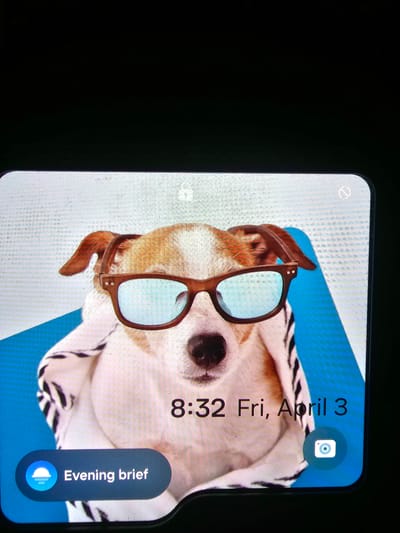 The image depicts a smartphone screen displaying a lock screen. The wallpaper is a picture of a dog wearing sunglasses and a hooded garment against a blue and white background. The lock screen displays the time as 8:32, the date as Friday, April 3, a camera shortcut icon, and a notification labeled "Evening brief." The phone has a "do not disturb" icon in the upper left corner. The environment surrounding the phone is dark. There is no information in the image to identify the city of Abraka, Nigeria.