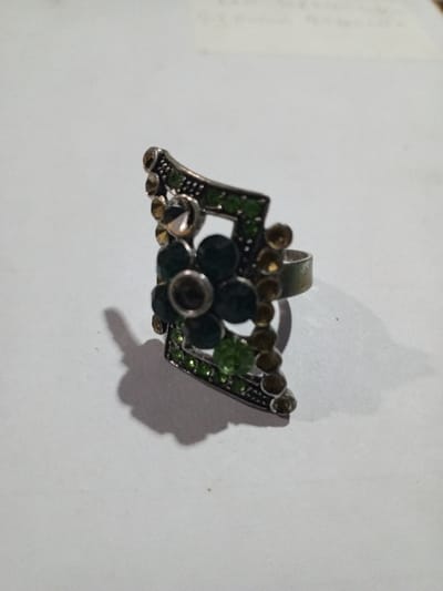 The scene, located in the city of Phakeo, Laos, depicts a single metallic ring resting on a light grey or off-white, planar surface. The ring features an elongated, shield-like design with a dark metal finish. Its face is adorned with multiple faceted stones: a central cluster forms a floral pattern using five dark green, circular stones, surmounted by a single larger, champagne-colored faceted stone. The outer perimeter of the ring's face is outlined with a row of small, bright green faceted stones, which is then bordered by a secondary row of small, amber-toned faceted stones. The ring's band is visible on the right side. A distinct, dark shadow is cast by the ring onto the surface, extending towards the lower left, indicating a light source from the upper right. No individuals, actions, or additional objects are present in the immediate vicinity.