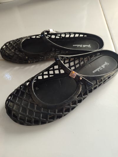 A pair of black, lattice-patterned jelly flats are placed on a white tiled surface. The shoes are positioned diagonally, with the front of the left shoe angled towards the bottom left of the frame, and the front of the right shoe angled towards the bottom right. Both shoes appear to be of a similar size. A strap with a small, metallic buckle crosses over the top of each shoe. The interior of the right shoe shows the brand name "Youth Balance" printed in white script. A size indicator, "38", is also visible on the insole of the right shoe. The tiled surface has visible grout lines running horizontally and vertically. The lighting suggests an indoor environment, with some natural light contributing to the illumination. There are no people or animals present in the image. The overall scene is still and appears to be a product photograph or a personal item display. The location is specified as Lancokpanteara, Indonesia.