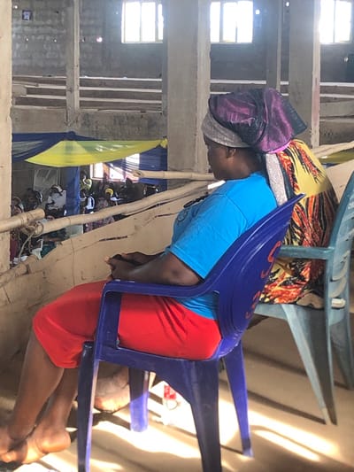 An adult in Nteje, Nigeria, wearing a purple head covering, a light blue t-shirt, and red shorts is partially seated in a blue plastic chair. The scene appears to be an indoor gathering of people seated on mats. In the background, structural elements of a building are visible, along with yellow and blue drapery. A partially visible window is on the upper-left. Other chairs are present in the foreground. The image is rotated 90 degrees clockwise.