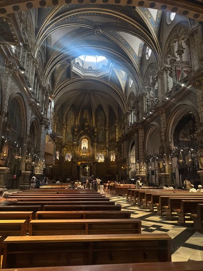 This is an interior view of a grand church, likely a cathedral or basilica, showcasing its ornate architecture and a congregation. The image is dominated by the high, arched ceilings, adorned with intricate gold detailing and painted murals. Natural light streams in from a large skylight or window at the apex of the nave, creating visible light shafts. Rows of wooden pews fill the central aisle, with a checkered floor pattern beneath. Several people are seated on the pews, while others are standing and milling about, appearing to be tourists or visitors. The altar area at the far end of the church is brightly lit and features religious iconography and a prominent cross. The overall impression is one of solemnity and architectural splendor. The church is identified as Santa Maria de Montserrat, located in the municipality of Monistrol de Montserrat, not Vacarisses, Spain.