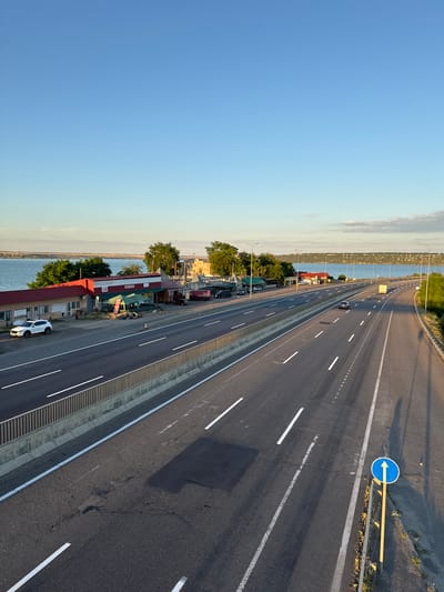 This is an aerial, wide-angle, eye-level shot of a highway passing by a body of water, presumably the Dnipro River, with a small commercial strip on the left side of the road. The sky is clear blue with a hint of yellow near the horizon. The highway has multiple lanes and is mostly empty, with a few cars and trucks visible. The commercial strip consists of several low-rise buildings with red roofs, one of which appears to be a cafe. The overall scene is calm and serene, with the warm golden light of what appears to be late afternoon or early morning casting long shadows across the road. There are no people visible in the image. The location is stated to be Yelyzavetivka, Ukraine.