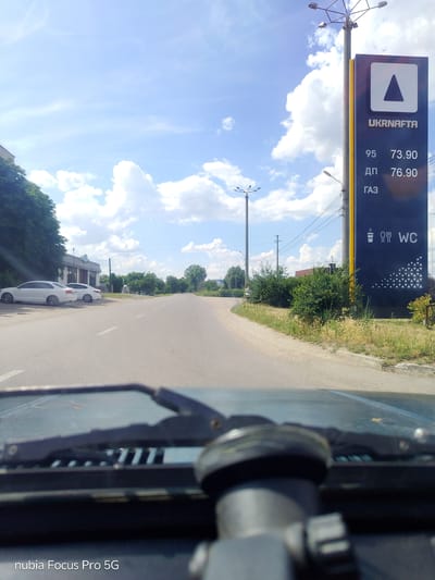 The image displays a gas station sign for UKRNAFTA, indicating fuel prices of 73.90 for 95 octane, 76.90 for diesel (ДП), and also listing GAS (ГАЗ). The sign also features icons for a coffee cup, cutlery, and a WC. The scene appears to be a roadside view, possibly from the perspective of a vehicle's dashboard, as the windshield wipers and a portion of the car's interior are visible in the foreground. Two white cars are parked on the left side of the road. The environment is sunny with a blue sky and scattered white clouds. The location is not Rímac, Peru; it is a street scene in a Ukrainian-speaking region, as indicated by the language on the sign.