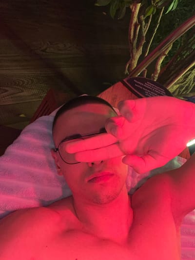 A male individual is depicted in a close-up, top-down perspective, lying on his back with his upper torso bare. He possesses short hair and wears thin-framed eyeglasses. His right hand is raised, with fingers covering his eyes and upper face. The entire scene is illuminated by a dominant red light source, casting a strong crimson hue over the subject and immediate surroundings. He is resting on a light-colored, possibly white, towel. In the background, to the upper left, wooden paneling is visible. To the upper right, a green leafy plant with visible branches stands beside a dark-colored sign. The sign features instructional text in both Romanian and English, specifically stating: "SANITIZE YOUR BED BEFORE AND AFTER USE." This environment is consistent with a wellness facility, such as a solarium or red light therapy room, located in Săftica, Romania.