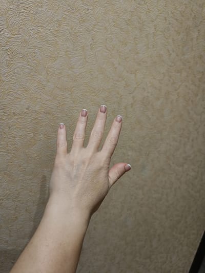 This media file features a close-up view of a person's left hand, held up against a textured, light brown wall. The hand is positioned with the palm facing away from the viewer and the fingers extended, with the thumb tucked in. The fingers display a French manicure, with neatly painted white tips. The skin on the hand appears fair with visible veins and some subtle lines, suggesting an adult. The background wall has a raised, organic pattern resembling dried leaves or brushstrokes, in a muted tan color. The lighting is even, and there are no strong shadows, indicating an indoor setting or an overcast day. No text is visible in the image. The overall impression is simple and focused on the detail of the hand and its presentation against the textured backdrop.