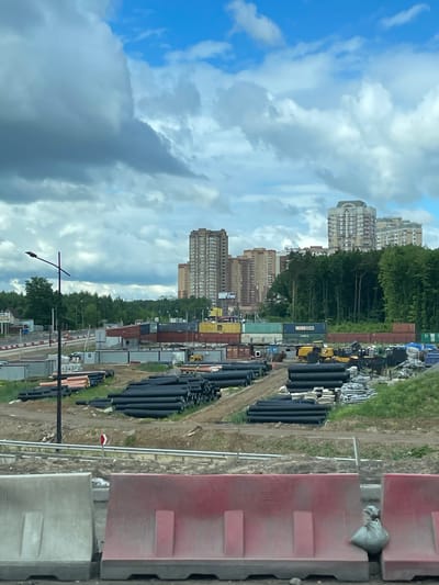 The image shows a view in Novodrozhzhino, Russia. The scene includes residential high-rise buildings, trees, stacks of black plastic pipes, a row of shipping containers and temporary office units, and construction barriers. The sky is cloudy with patches of blue. A lamppost is visible. The overall impression is of an urban environment with construction activity or material storage near residential areas.