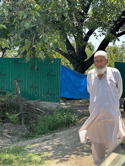 In the city of Vessu, India, an elderly man with a white beard and a white skullcap stands in the foreground. He is wearing a light-colored, loose-fitting tunic and pants. Behind him, a large tree with green leaves provides shade. A bright blue tarp is visible behind the man and to his left, and a green corrugated metal fence is in the background. The ground is dirt and gravel, with some green vegetation growing on the left side of the image. The lighting suggests it is daytime.