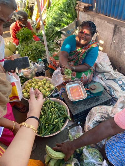 In Chinnambedu, India, a bustling market scene unfolds. A woman, her face illuminated by sunlight, sits behind a vibrant display of fresh produce. She wears a blue top and a colorful sari adorned with floral patterns, her hair tied back and a red bindi adorning her forehead. To her right, a traditional weighing scale sits ready for use, with a brass weight resting on it. In front of her, a pile of okra fills a metal bowl, while a larger bowl nearby holds a bounty of small, green eggplants. A man in a red shirt, his face partially obscured by a large bunch of leafy greens, appears to be engaged in selling produce. Shoppers, their hands reaching out, are visible in the foreground, examining or purchasing vegetables. The market floor is littered with sacks and bags, indicative of a busy marketplace. The overall atmosphere is one of commerce and community, with a focus on fresh, locally sourced produce.