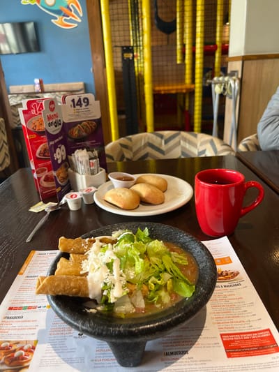 The table is set with a red mug, a white plate of fried foods with a small dipping container, and a black bowl of food with lettuce and white toppings. The table also contains a menu with prices and descriptions of food items, including a daily menu for $14.99 and another for $99. There are also small condiment containers and a silver spoon. A children's play area with yellow bars is visible in the background.