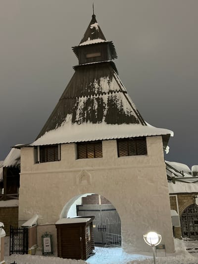 The image displays a snow-covered architectural structure at night, located in Matiushino, Russia. The central subject is a robust building, possibly a gatehouse, constructed with white, textured plaster walls and topped by a prominent, dark wooden conical roof. The roof features several tiers of dark wooden shingles, heavily blanketed with thick white snow along its eaves and sloped surfaces. The apex culminates in a smaller, belfry-like structure with a rectangular opening and a pointed finial.

Below the wooden roof, the white plastered facade contains three rectangular windows with dark frames and internal grid patterns, emitting a warm, artificial light from within. A large, rounded archway forms a passage through the base of the building at ground level. The ground, both leading to and within the archway, is covered in a substantial layer of pristine white snow.

To the left of the archway, a section of a black wrought-iron fence is partially visible amidst the snow. Adjacent to this fence stands a small, dark wooden kiosk or booth with horizontal slat siding, also partially obscured by snow. A white sign with dark text and a directional arrow is positioned near the kiosk. Inside the archway, a large double-leaf wooden gate with dark metal hardware is visible in the background, alongside a white metal barrier to its right. To the right of the archway, a modern street lamp featuring a spherical white diffuser on a dark pole illuminates the immediate foreground. Further right, a segment of an adjoining snow-covered building with an arched window is present. The sky above is uniformly dark, indicating nighttime conditions. No people, specific actions, or interactions are visible within the scene.