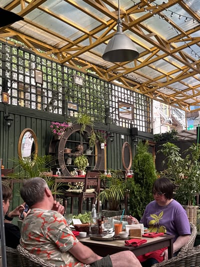 The image depicts an outdoor seating area, possibly a cafe or restaurant, with a covered roof structure. The roof is supported by a wooden frame and covered with a translucent material, allowing diffused light to enter. String lights are suspended under the roof. A large industrial-style metal pendant lamp hangs from the center of the roof. The sides of the seating area are enclosed by dark green wooden panels and a black lattice screen, through which foliage can be seen.

In the foreground, a man with gray hair wearing a camouflage t-shirt with orange accents is seated at a table. He is looking to his right, with his left hand touching his chin. He is holding a mobile phone in his right hand. To his left, another person is partially visible, also looking at a phone.

To the right, a woman with dark hair and glasses is seated at the same table. She is wearing a purple t-shirt with a yellow fruit design and is holding a fork with a piece of food, seemingly eating. A glass of orange liquid, possibly juice, and a white napkin are on the table in front of her. Red napkins and cutlery are also arranged on the table.

Behind the table, a large, dark, circular decorative clock face with Roman numerals is mounted on the wall. It features shelves with various items, including potted plants, decorative pots, and a bell. Several potted plants are placed around the seating area, adding to the lush greenery. In the background, glimpses of buildings with brick facades and windows are visible through the lattice and roof structure. The overall scene suggests a relaxed outdoor dining environment.

While the prompt mentions the city of Zhukovka, Russia, there are no specific visual cues within the image that can definitively confirm this location.