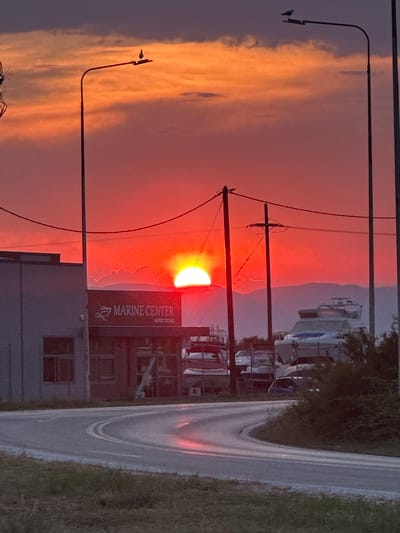 An extreme wide shot captures a vibrant sunset over the city of Chaidefto, Greece. The sun, a brilliant orange orb, is partially obscured by a layer of clouds and mountains on the horizon. The sky is a dramatic gradient of red, orange, and purple hues, with scattered clouds reflecting the sunset's intensity. In the foreground, a winding asphalt road curves through the scene, reflecting the warm light. On the left side of the road, a modern, grey building with windows and a sign that reads "MARINE CENTER" stands prominently. Several boats are visible behind the building and further to the right, suggesting a marina or boat storage area. Two tall streetlights with birds perched on them stand on either side of the road, framing the sunset. The environment appears to be an open area, possibly a coastal road leading into or out of a town. The overall impression is one of a serene and picturesque evening.