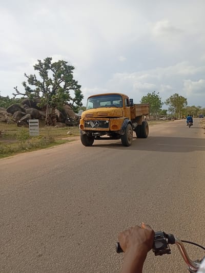 The image shows a yellow truck driving on a paved road in Shuwa, Nigeria. The truck is old and appears to be used for hauling. In the background, there are trees, rocks, and buildings. A person's hand is visible on the handlebars of a bicycle or motorcycle in the foreground, suggesting the photo was taken from a moving vehicle. The sky is cloudy.